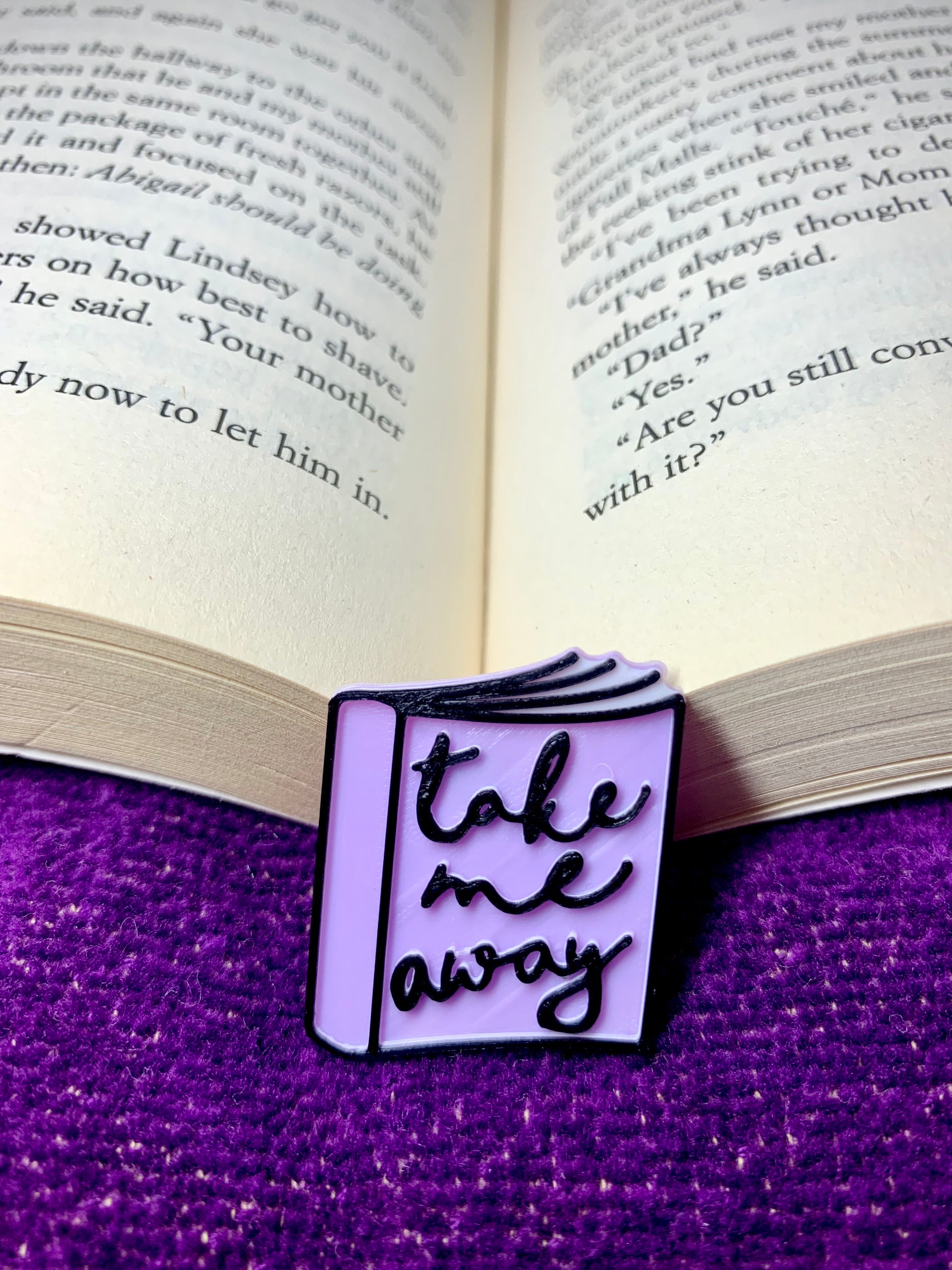 Resting in the spine of a book and resting on a purple beach tile is a 3D printed pin from R+D. The pin looks like a paperback book with white pages and a light purple spine. Details on the book are accentuated with black eco friendly filament. On the cover, it reads, take me away in a cursive script. 