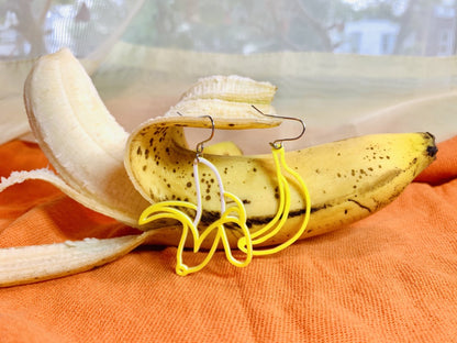 Resting on a orange cloth napkin is a half peeled ripe banana. Two R+D earrings are hanging off of the peel. One is an unopened, unpeeled yellow banana. The other is a half peeled banana that reveals a white banana inside.