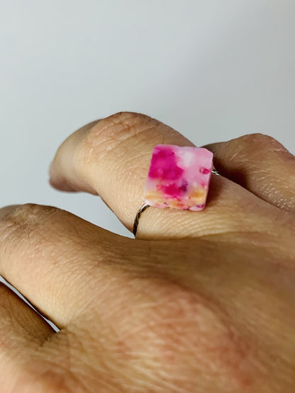 There is a hand on a white background wearing a cast ring in the shape of a square. The ring is cast from recycled 3D prints in warm hues of pink, yellow, white, red, orange and purple. It has the effect of granite or a speckled egg. 