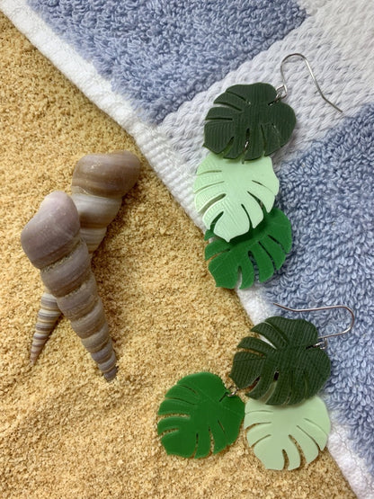 There are two earrings laying on a blue and white striped beach towel. They are small monster leaves that are layered together. The leaves are each different gradients of green: olive green, mint green, and kelly green. The towel is laying over bright fully sand with two twisted shells sitting nearby.
