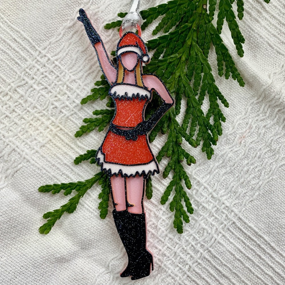 On a white fabric background there is a green branch from a tree. Laying on top of these is a 3D printed ornament from R+D. The ornament is printed in a plant based filament. It is shaped like Regina George from the movie Mean Girls. She is striking the iconic pose at the beginning of performing Jingle Bell Rock, wearing black gloves and boots and a red outfit with white trim. 