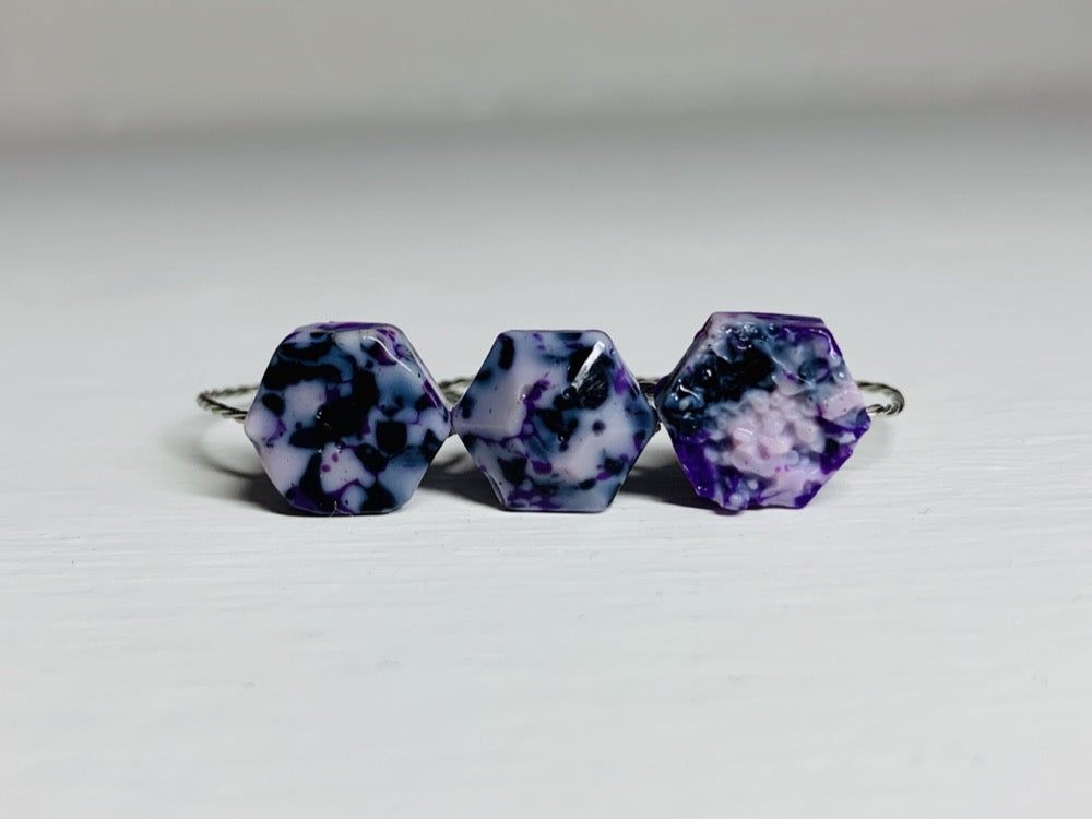 There are three cast rings sitting side by side. They are each in hexagon shapes: one smooth, one faceted, and one with the texture of a geode. They are cast from recycled 3D prints in hues of purple, black and white. 