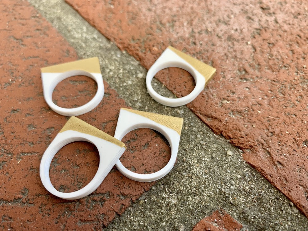 Laying across a brick pathway are four 3D printed rings. They are each smooth circle bands that extend into a square shape above the finger where worn. They are printed in a white plant based filament and dipped into a gold paint to give a unique addition across the top of each ring.