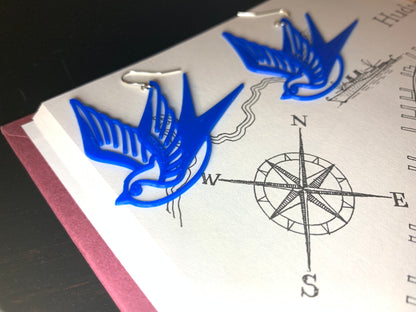 Laying on a book with a hand drawn compass are two bright cobalt blue swallow R+D 3D printed earrings. The shape of these birds is reminiscent of classic sailor tattoos. 
