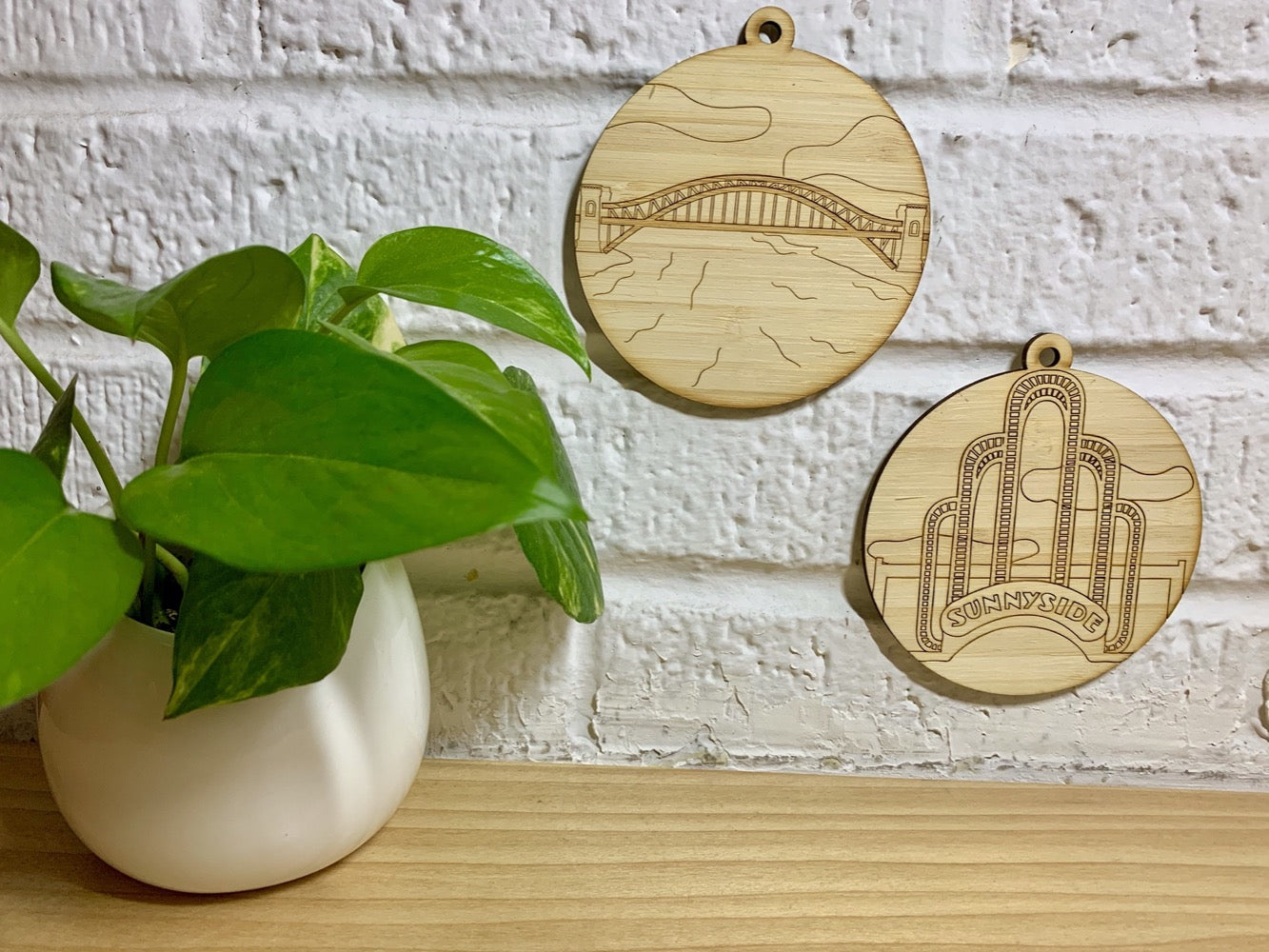 Shown on a white brick wall and next to a houseplant are two laser cut wall hangings. It is cut from bamboo, a fast growing and sustainable grass. Etched in one is a picture of the iconic Sunnyside Arch in the center of the Queens neighborhood. The other shows the Hell Gate Bridge in Astoria. Each piece can be painted like a paint by number or left plain. It can hang from your Christmas Tree or on you wall all year round.