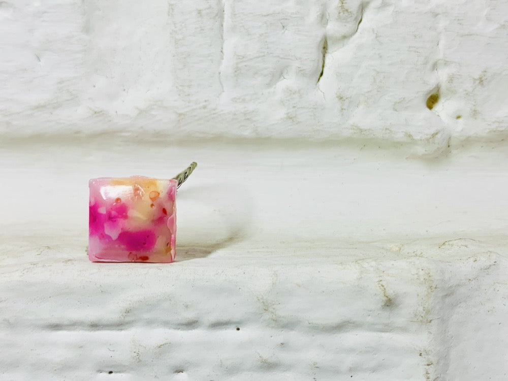 Resting on white painted bricks is a ring that is cast in the shape of a faceted square. The ring is cast from recycled plant based 3D prints. in pinks, reds, yellows, oranges, and white. The colors are mixed together to give a speckled or marbleized appearance. 