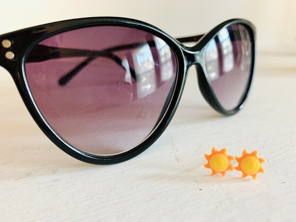 In the foreground are two small stud earrings. They are shaped as suns with bright yellow centers that are rounded and eight sun rays that curl around the sun in orange. In the background are a pair of black sunglasses.