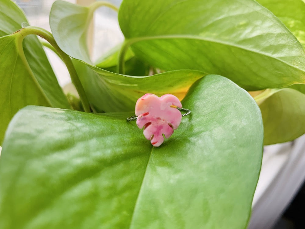 On bright green leaves is a ring with a cast charm resting. The ring is in the shape of a monstera leaf. It is cast from recycled plant based 3D prints in light pink, hot pink, white, red, yellow, and orange.
