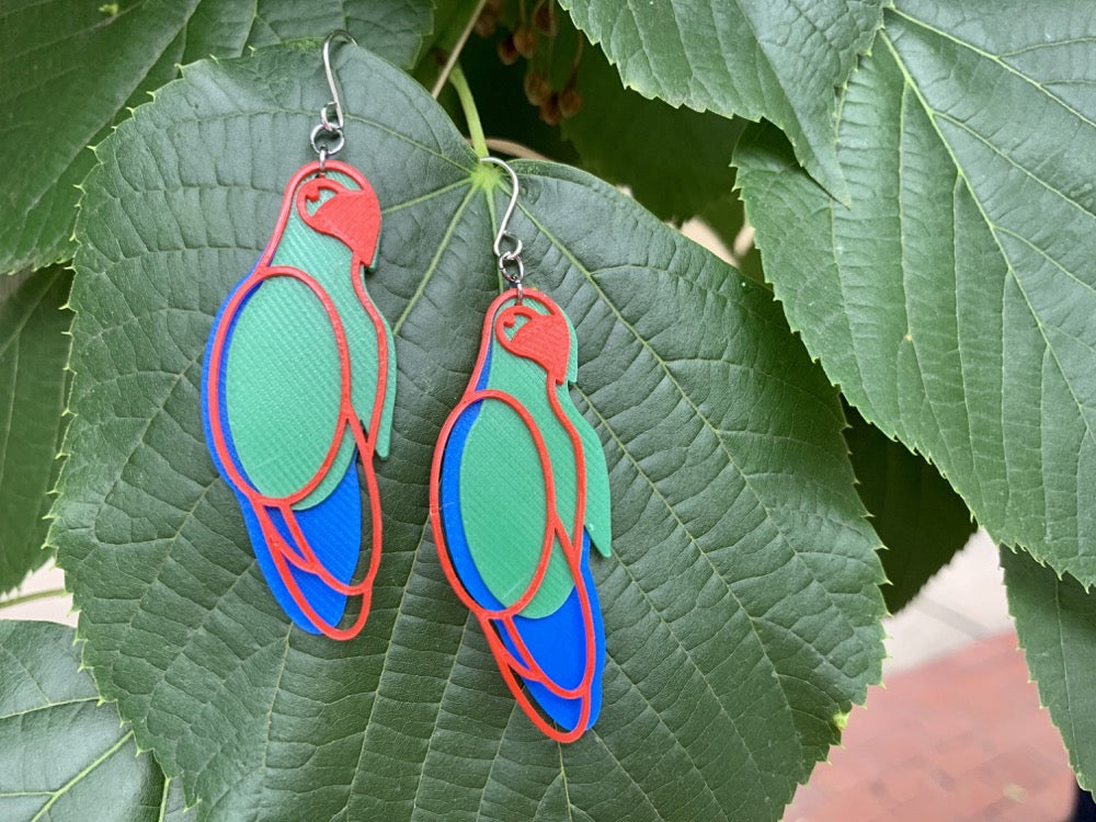 Hanging off of a large lush green leaf are two R+D earrings. The earrings are made from three pieces , one cobalt blue, one kelly green, and the final, which rests on top of the other is bright red and creates the outline of a parrot. 