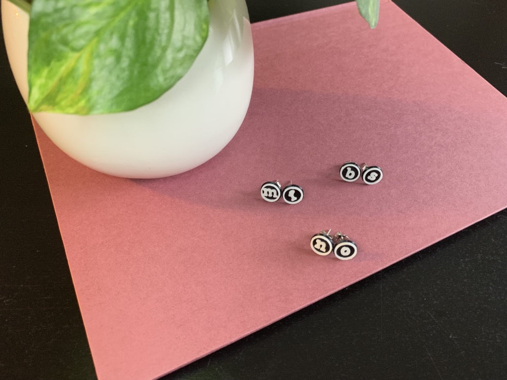 There are three pairs of earrings shown. These r+d earrings are shaped like typewriter studs, with black backgrounds, white circle outlines and a lowercase letter in the center. They are 3d pritned with an eco friendly and plant based material. The earring pairs are "bs", "mf", and " no". They are resting on a muted red book with a white ceramic planter next to it. 