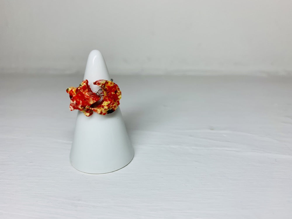 On a white background and slipped on a white cone ring holder are two rings that is cast from recycled 3D prints. The rings are in the shapes of a star and a moon with red, yellow, orange, and black colors intermixed to be speckled like granite.