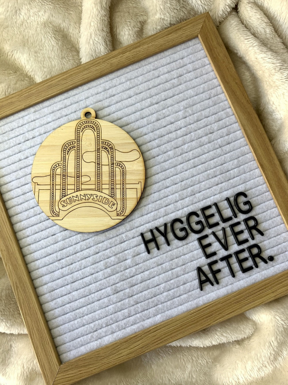 Shown on a cozy blanket is a letter board that reads "Hyggelig Ever After." There is also a laser cut wall hanging. It is cut from bamboo, a fast growing and sustainable grass. Etched in it a picture of the iconic Sunnyside Arch in the center of the Queens neighborhood. Each piece can be painted like a paint by number or left plain. It can hang from your Christmas Tree or on you wall all year round.