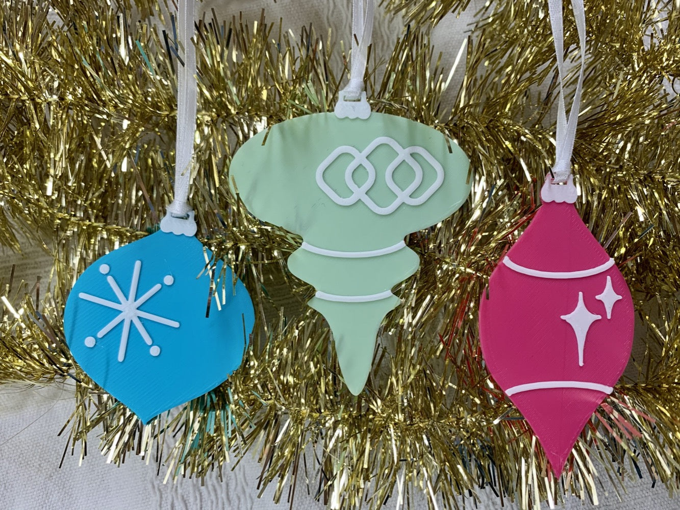 On a gold tinsel garland background are a set of 3 R+D 3D printed ornaments. They are all designed to look like vintage baubles that would have commonly been found on Christmas trees in the past. They each have white accents of stars and shapes. One is hot pink, one is mint green, and one is teal.