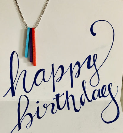 Hanging in a card with the words, happy birthday scrawled, is a necklace with three pendants. They are long rectangles in teal, purple, and red. When turned to the side, each pendant reveals a name. 