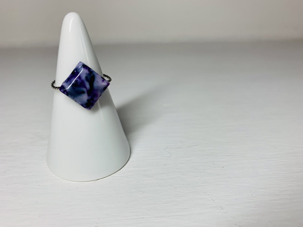 On a white background is a ring holder with a single cast ring resting on it. The ring is a square faceted shape with a twisted metal band. The ring is cast from recycled 3D prints in shades of purple, black and white to create a marbled or granite like appearance. 
