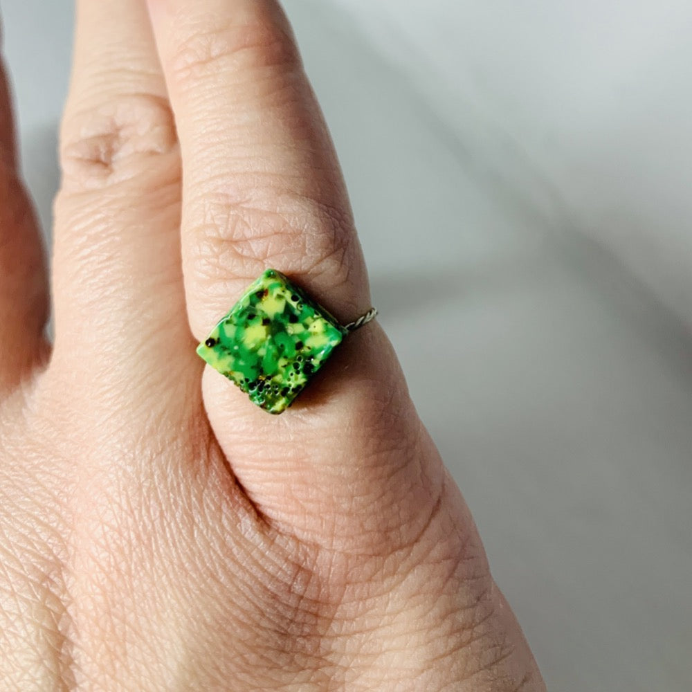 This is a close up of a person's pinky finger. They are wearing a ring that is a square shaped charm with a thin twisted metal band. The charm is made from recycled 3D prints in hues of green. 