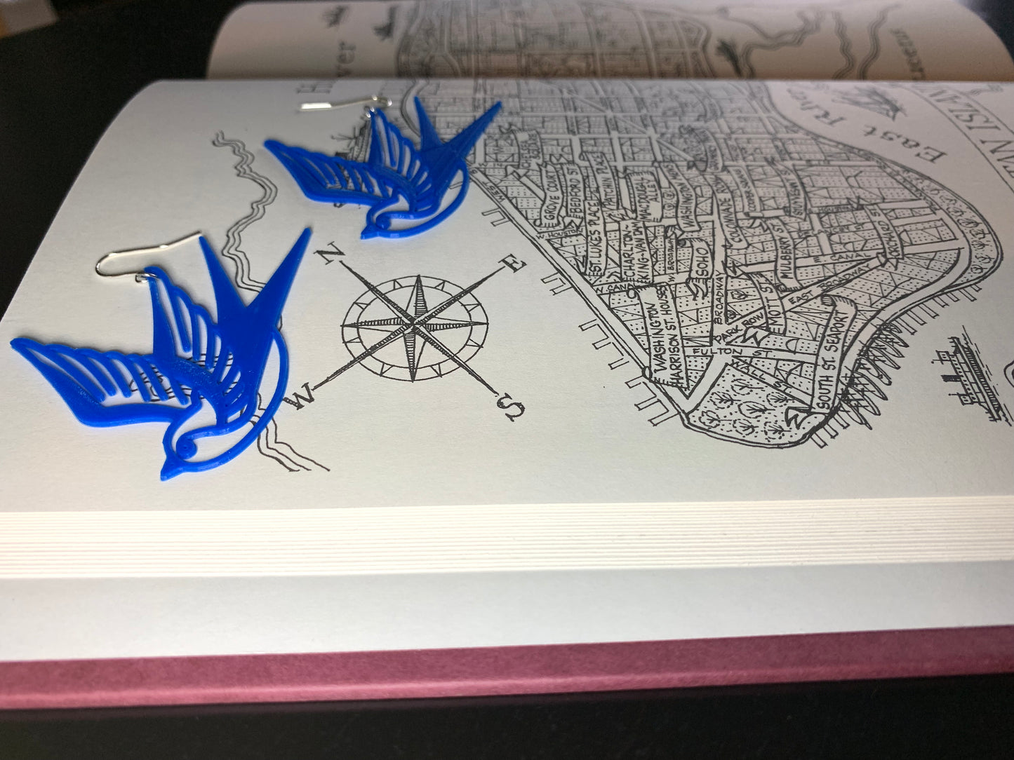 Laying on a book with a hand drawn compass are two bright cobalt blue swallow R+D 3D printed earrings. The shape of these birds is reminiscent of classic sailor tattoos.