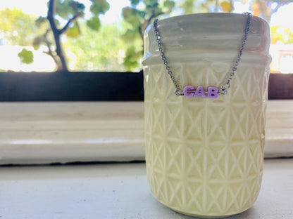 Hanging off of a ceramic mason jar is a necklace with small letters on a metal bar. The letters are like beads and will rotate and move around. This necklace has 3 characters (CAB) in a light purple. 