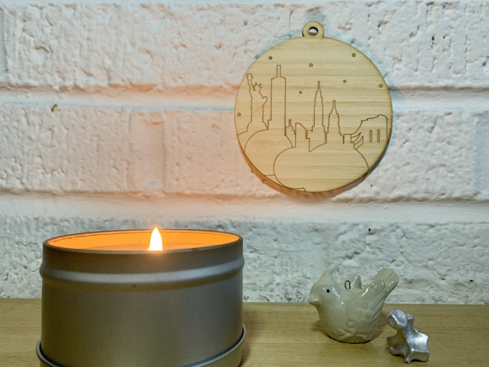 Shown on a white brick wall next to a candle and the knick knacks, is a laser cut wall hanging. It is cut from bamboo, a fast growing and sustainable grass. Etched in it a picture of the New York City Skyline with the Statue of Liberty, Empire State Building, Chrysler Building and Brooklyn Bridge all visible. Each piece can be painted like a paint by number or left plain. It can hang from your Christmas Tree or on you wall all year round.