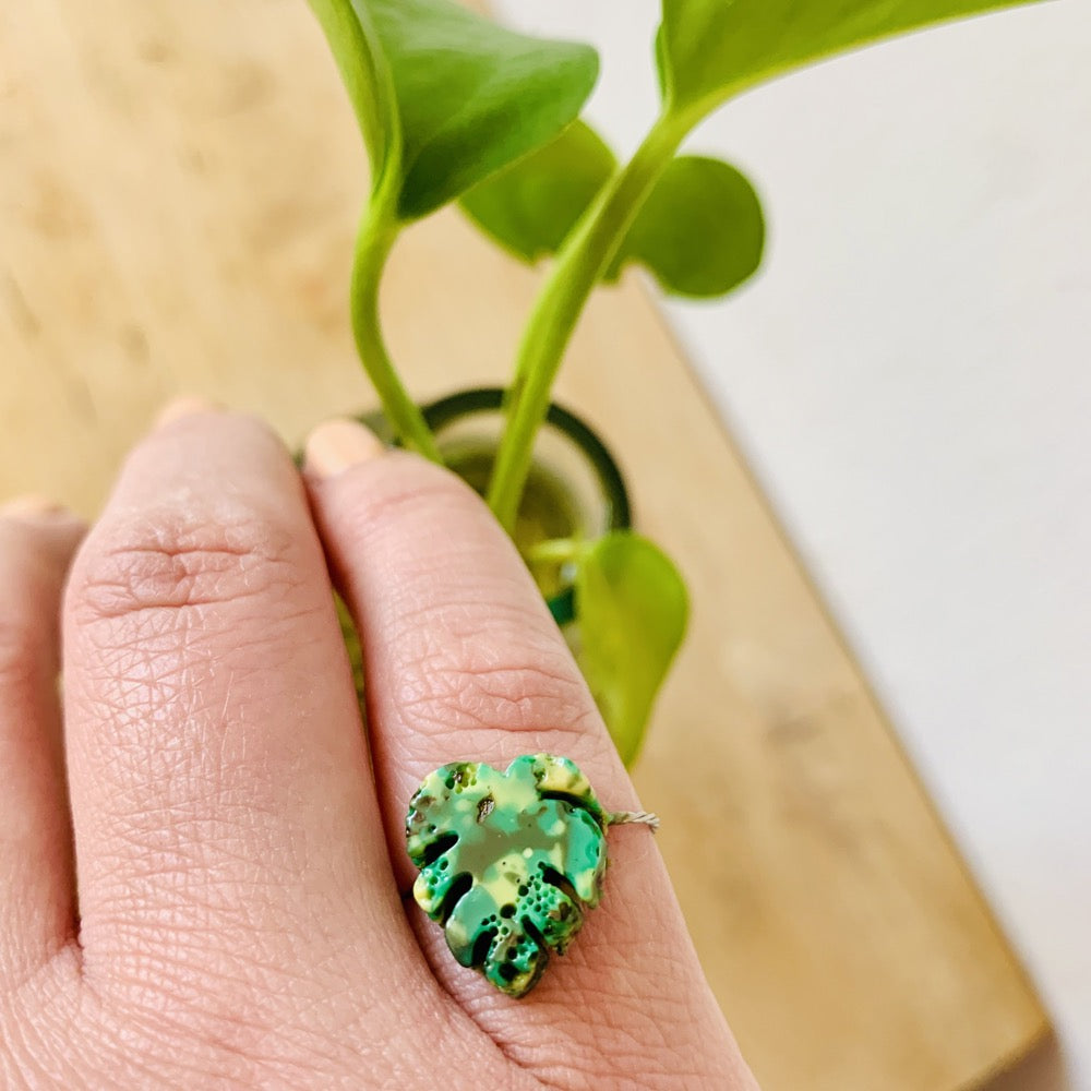On a persons pinky finger is a green ring cast from recycled 3D prints. It is in the shape of a monstera leaf and made from kelly green, light green, and olive green filaments. 