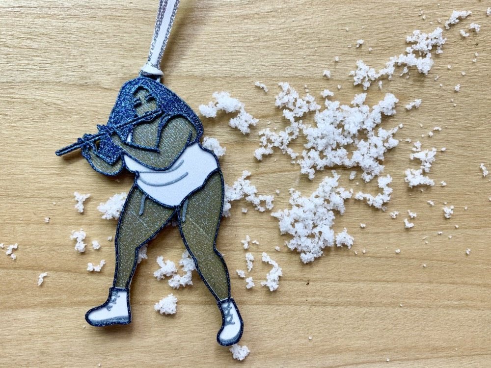 On a light wood shelf is a scattering of snow. Resting on top of this is a R+D 3D printed ornament. The ornament is shaped like the singer, rapper, and musician Lizzo. She is wearing a white leotard, bend slightly, and playing her flute name Sasha Flute. The entire ornament is printed with a plant based filament and covered in glitter to give it a perfect shimmer and shine.