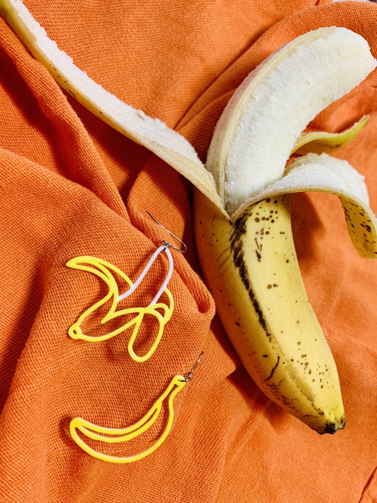On an orange cloth napkin there is a ripe banana that is halfway peeled. Next to it are two R+D 3D printed earrings. One is a half peeled yellow banana that reveals a white banana. The other is a fully unpeeled banana. 