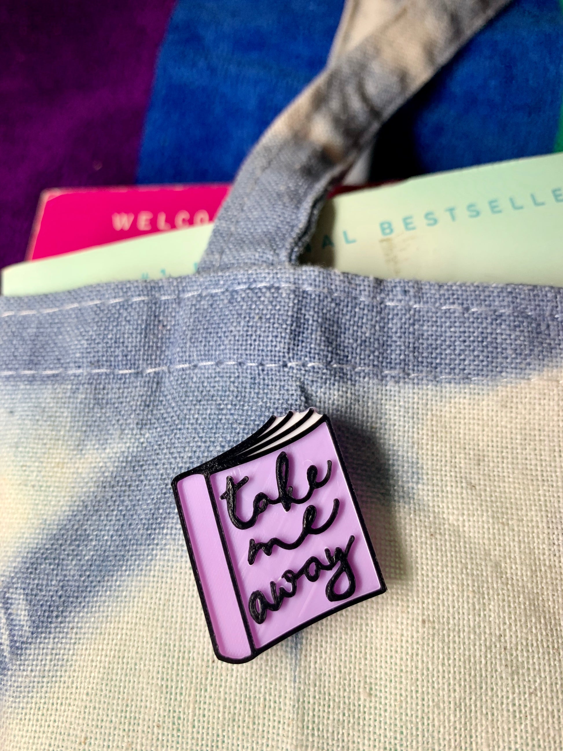 There is a close up of a cotton tote bag with a blue and white shibori pattern. Spilling out of the bag are a couple of paperback books suitable for reading on the beach. Attached to the tote bag is a 3D printed pin shaped like a floppy well read book. The pin is light purple with white pages and black details. On the cover ar scripted cursive words that say take me away. 