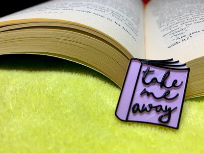 Resting on a bright green beach towel is an open paperback book. In the spine is a small 3D printed pin from R+D. The pin is light purple and shaped like a floppy paper back book. There are white pages and black scripted letters that say, take me away. 