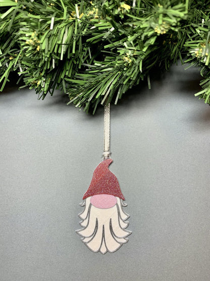 On a grey background and hanging from a green wreath is a R+D 3D printed ornament. This ornament is shaped like a gnome, elf, or plump santa. It has a red pointed hat, a big pink nose, and a white and silver beard. The entire ornament is covered in glitter to be able to shimmer and shine in the light.
