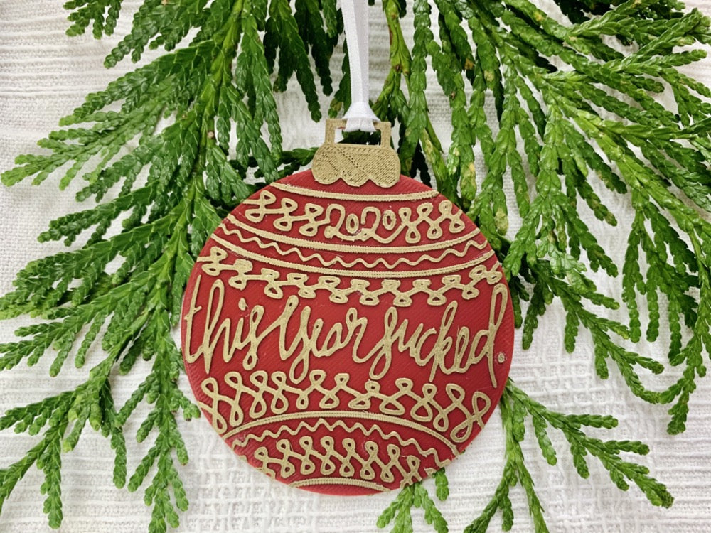 On a white fabric background there are sprigs of everygreen and a R+D 3D printed ornament. The ornament is shaped like a traditional bulb. It is dark red with gold drawings and script on it. Worked into the doodles are the year 2020 and then the words, "this year sucked".