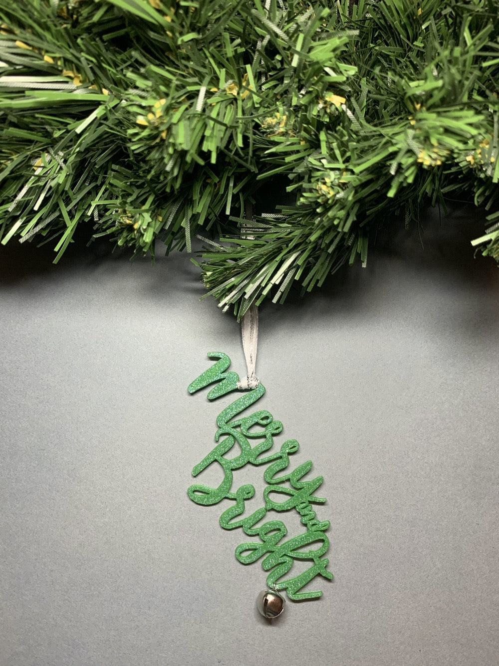 On a grey background and hanging from a green wreath is a 3D printed R+D ornament. It is a cursive text with a jingle bell and covered in glitter to make it shimmer and shine in the light. This ornament reads, Merry and Bright.