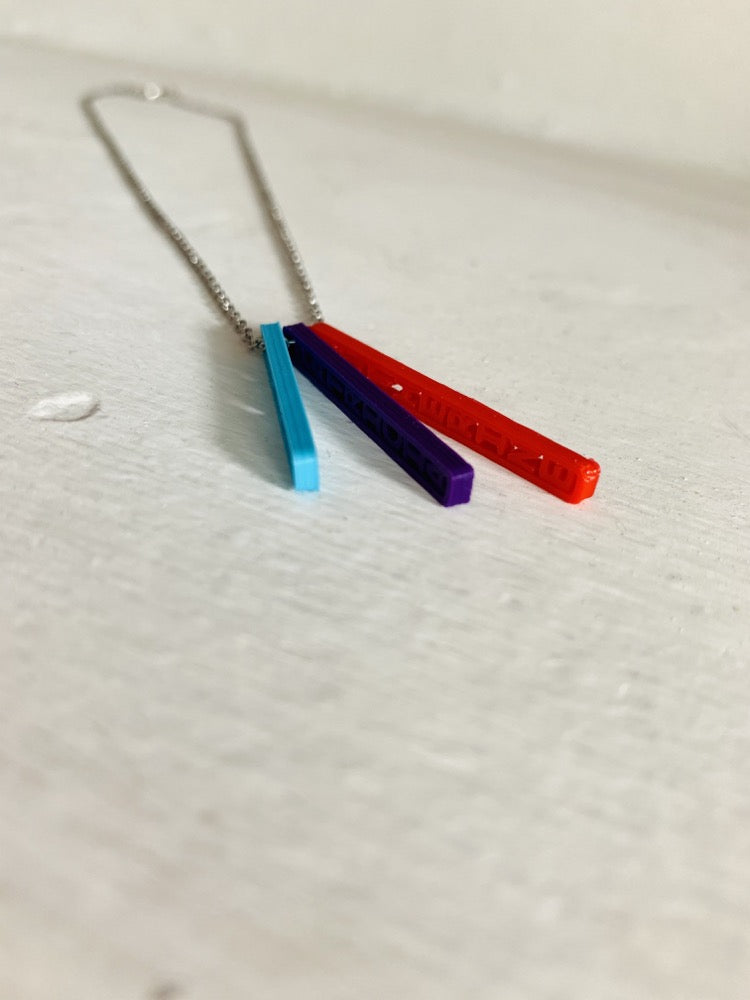 On a white background are three 3D printed pendants. They are each different colors and shaped as long rectangles with names in the center. When worn, they simply look like long hanging rectangles, but from the side, all the names are visible. This one has a teal pendant, a purple pendant, and a red pendant. 