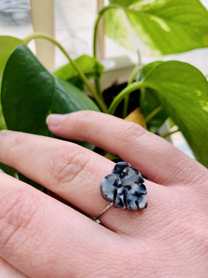 In the foreground is a hand with a ring cast from recycled plant based 3D prints. It is a ring in the shape of a monstera leaf with black, white, and silver filaments being used to create a speckled look like granite. In the background are bright green leaves.