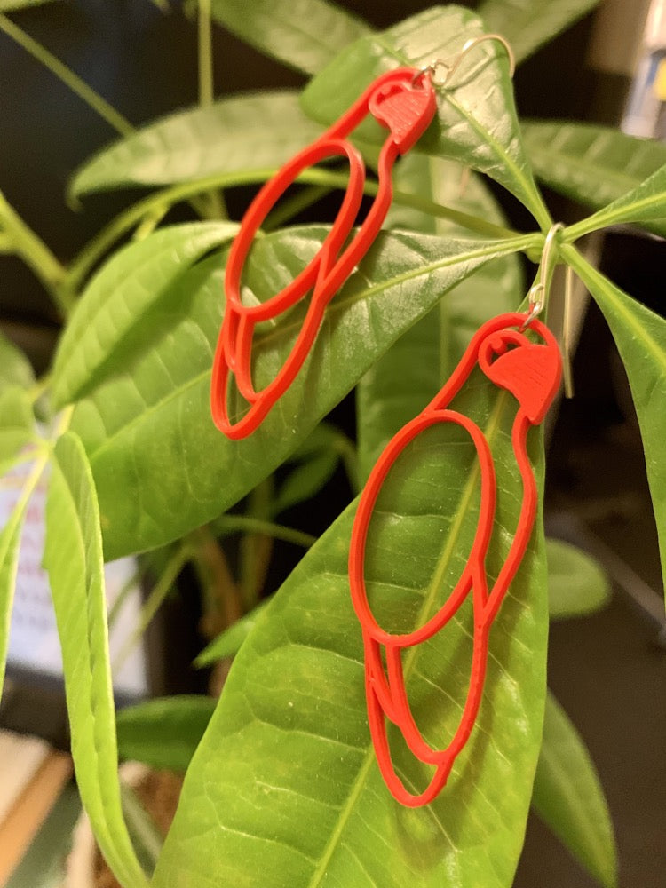 Two R+D earrings are shown hanging off of lush green leaves. The earrings are bright red and shaped like parrots. 