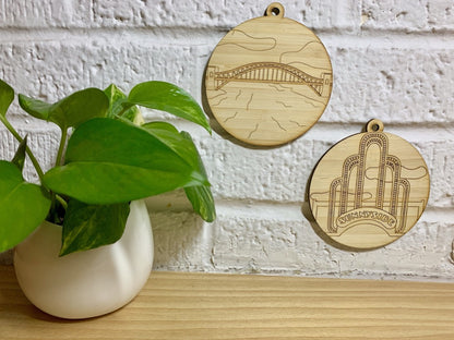 Hanging on a white brick wall and next to a houseplant are two laser cut wall hangings. They are each cut from bamboo, a fast growing and sustainable grass. Etched in one is a picture of the Hell Gate bridge in Astoria. This bridge is an icon of the area and looked on from Asttoria Park. The other features the sunnyside arch on Queens Blvd. These can be used as a ornament in the holidays or a wall hanging all year round. It can be painted like a paint by number or left plain.