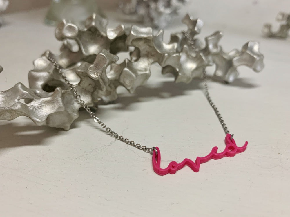 On a white background and with an aluminum sculpture, there is a necklace in the foreground. It is 3D printed and reads loved in a modern cursive script in a hot pink eco friendly filament.