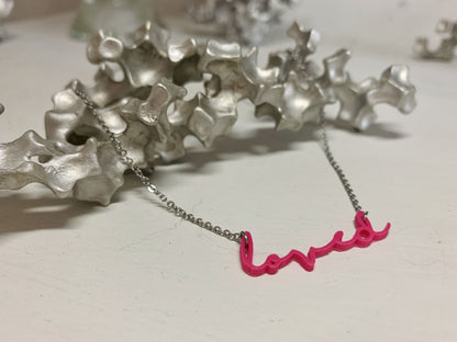 On a white background and with an aluminum sculpture, there is a necklace in the foreground. It is 3D printed and reads loved in a modern cursive script in a hot pink eco friendly filament.