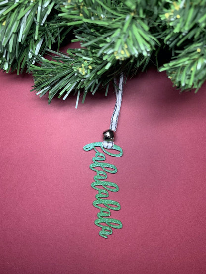 On a bright red background and hanging from a green wreath is a 3D printed R+D ornament. It is a cursive text with a jingle bell and covered in glitter to make it shimmer and shine in the light. This ornament reads, falalalala.