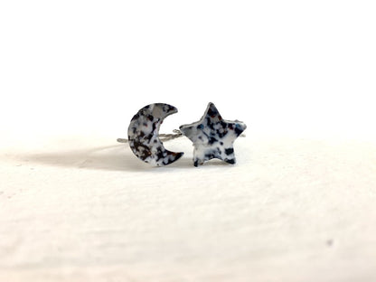 On a white background are two rings next to each other. One is a cresent moon shape, the other a star. They are cast from recycled 3D prints in black, white and silver plant based filaments. They have a speckled look like granite. 