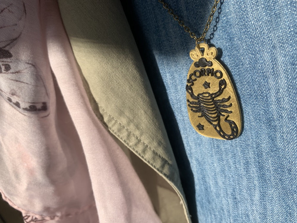 Hanging around someone's neck and peeking out from a jacket and scarf is an R+D necklace. The necklace is a gold zodiac pendant with black details. It is 3D printed with a plant based filament. This pendant has the sign Scorpio on it with a scorpion and stars.