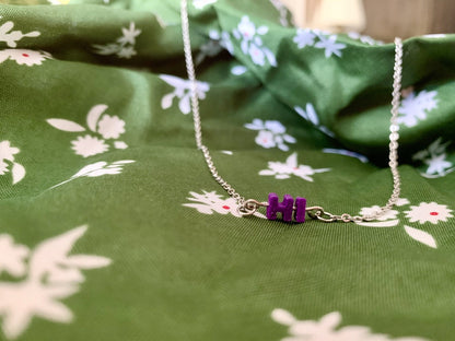 On green fabric with white flowers is a necklace with a metal bar. On the metal bar are two 3D printed letters, HI, in purple. 
