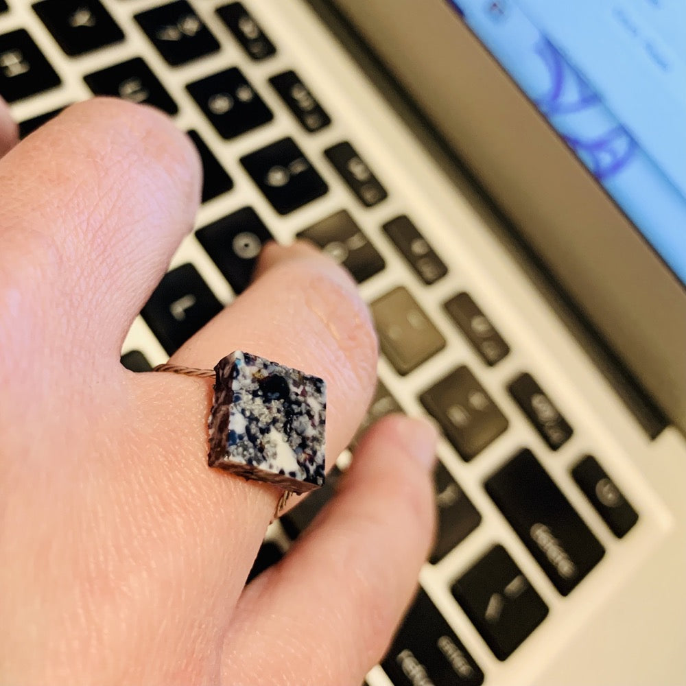 Shown on someone's finger as they type on a computer keyboard is a cast ring. The ring is a square shape with a smooth surface. It is made from recycled 3D prints that were black, white, and silver. It has a speckled look like granite. 