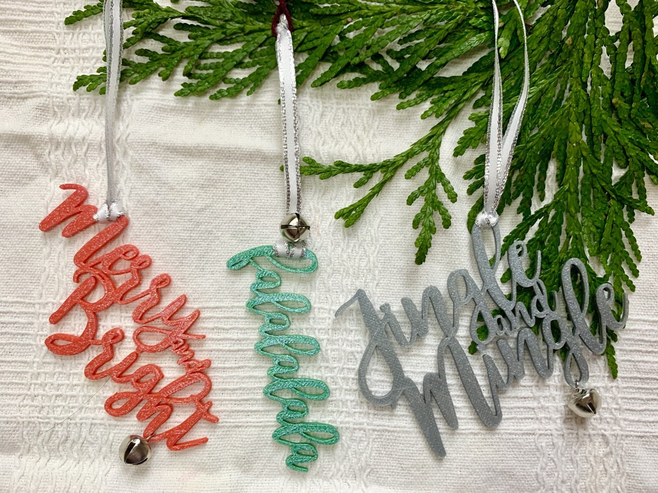 On a white fabric background there are three R+D 3D printed ornaments hanging from an evergreen branch. Each is in a cursive font and have a jingle bell attached. There are in red, green or silver colors and read "Merry and Bright", "falalalala", and "Jingle and Mingle"