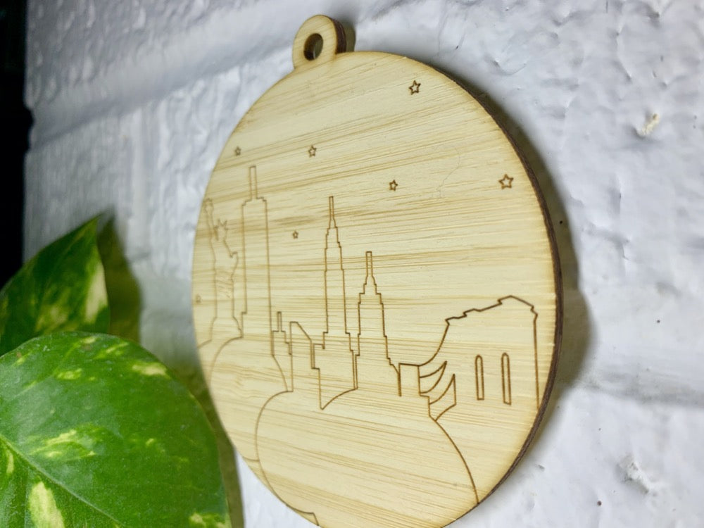Shown on a white brick wall and from the side so you can see the details is a laser cut wall hanging. It is cut from bamboo, a fast growing and sustainable grass. Etched in it a picture of the New York City Skyline with the Statue of Liberty, Empire State Building, Chrysler Building and Brooklyn Bridge all visible. Each piece can be painted like a paint by number or left plain. It can hang from your Christmas Tree or on you wall all year round.