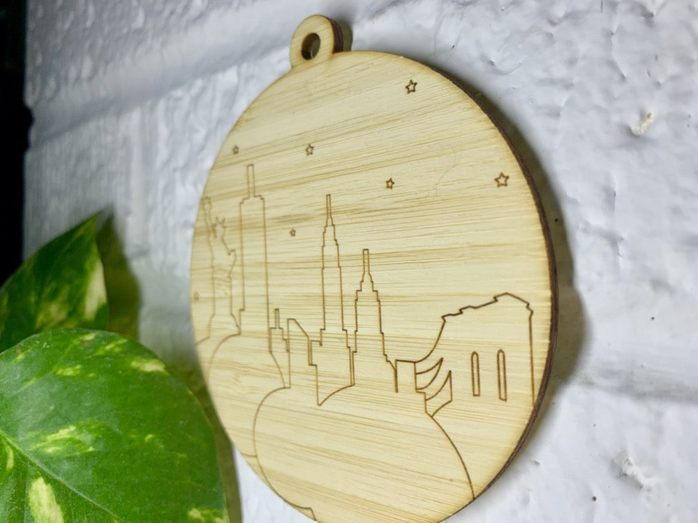 Shown on a white brick wall and from the side so you can see the details is a laser cut wall hanging. It is cut from bamboo, a fast growing and sustainable grass. Etched in it a picture of the New York City Skyline with the Statue of Liberty, Empire State Building, Chrysler Building and Brooklyn Bridge all visible. Each piece can be painted like a paint by number or left plain. It can hang from your Christmas Tree or on you wall all year round.