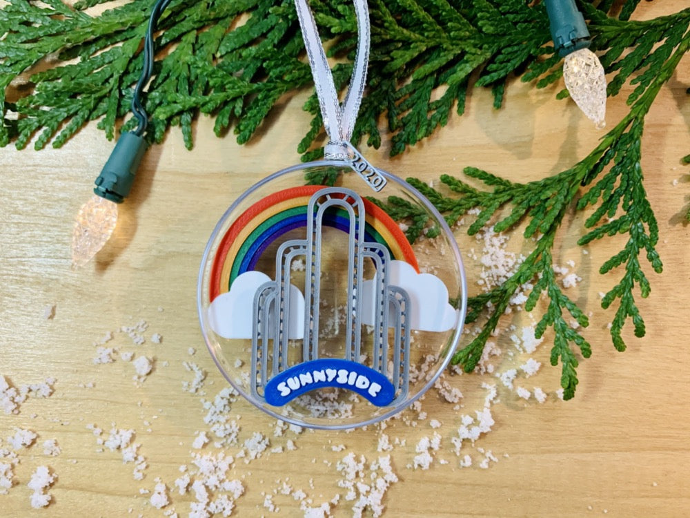 On a wooden shelf, there is a sprig of greenery, Christmas tree lights, and some scattered snow.Laying on top is a ornament with a small tag that has been 3D printed to read 2020. Inside the half circle ornament is a rainbow spanning between two clouds and the iconic Sunnyside Arch in front of it. 