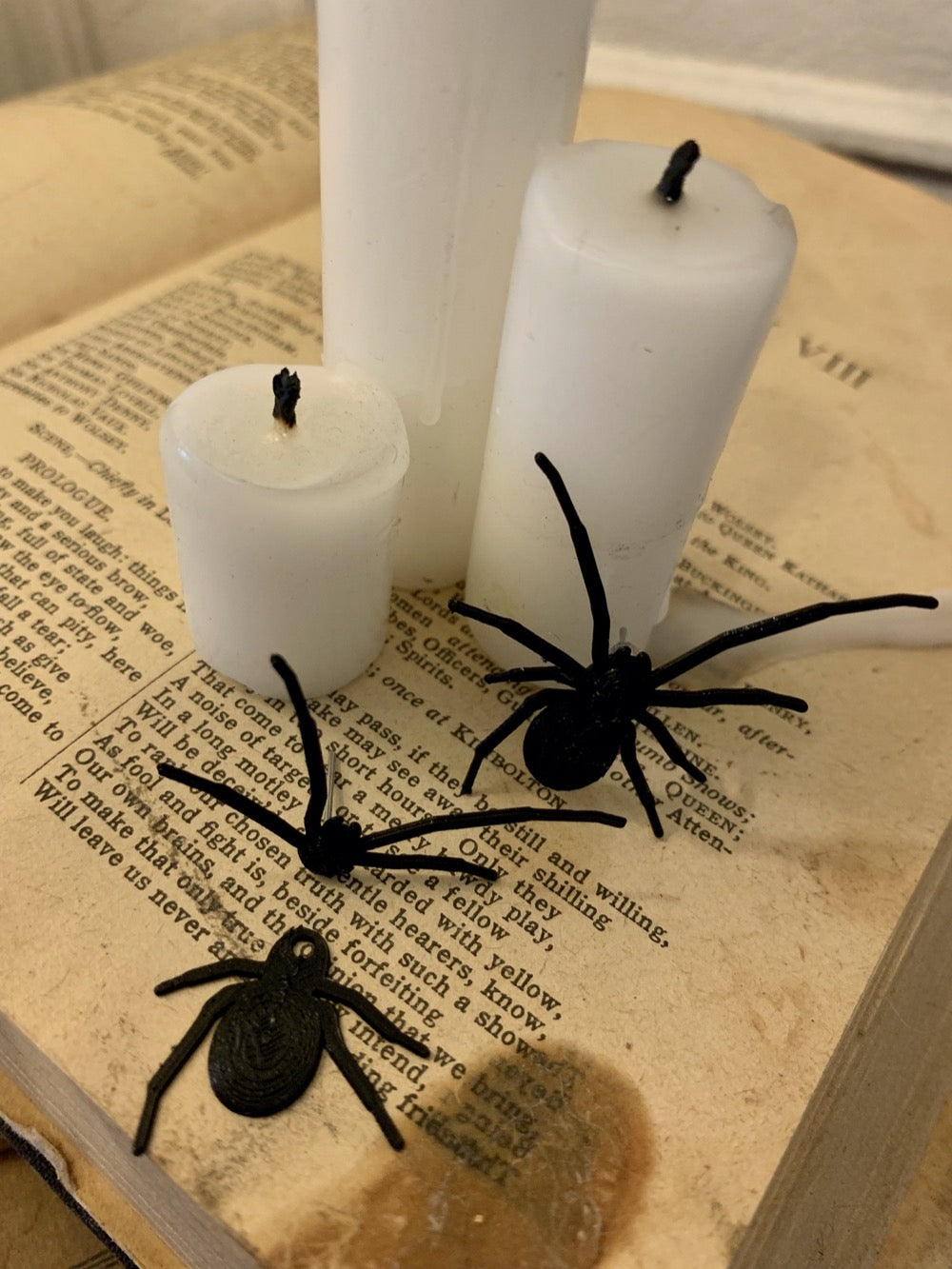 Shown are two black spider earrings that are realistic in their shape. They are worn as front and back earrings. One of the earrings has the two pieces separated to show how they would be then worn on each side of your earlobe. They are pictured crawling up an old book with candles on top to create a spooky Halloween scene.