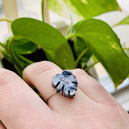 In the foreground is a hand with a ring cast from recycled plant based 3D prints.  It is a  ring in the shape of a monstera leaf with black, white, and silver filaments being used to create a speckled look like granite. In the background are bright green leaves. 
