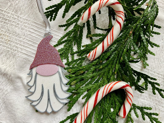 On a white fabric background there are evergreen branches and candy canes next to a R+D 3D printed ornament. This ornament is shaped like a gnome, elf, or plump santa. It has a red pointed hat, a big pink nose, and a white and silver beard. The entire ornament is covered in glitter to be able to shimmer and shine in the light.