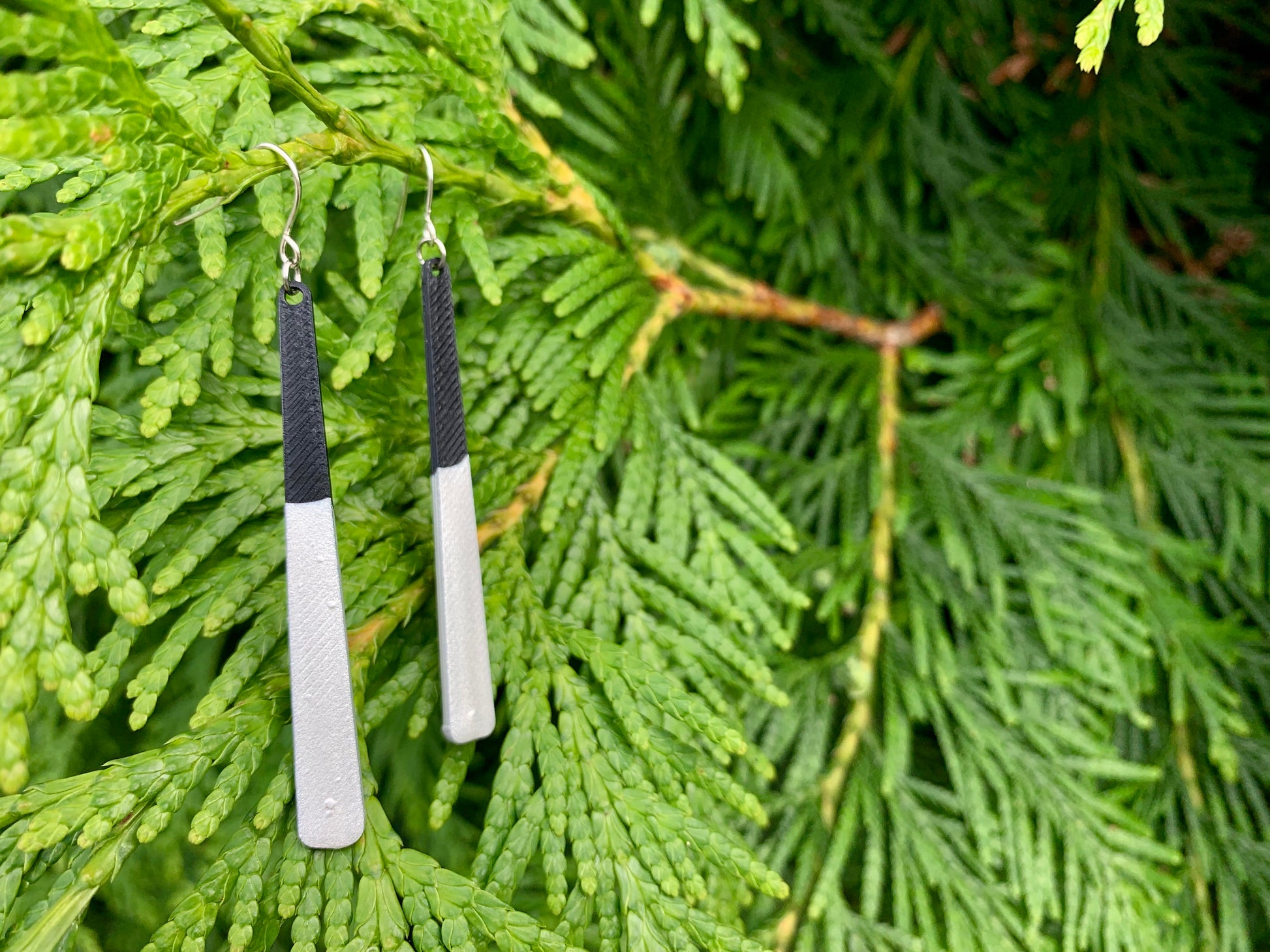 Two earrings are hanging off of lush green evergreen branches. They are long black dangles that have been dipped in silver metallic paint. 